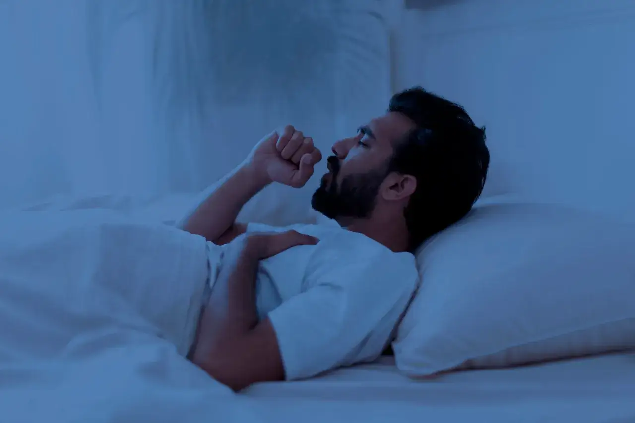 Un homme barbu, allong&eacute; dans son lit, se r&eacute;veille en pleine nuit pour tousser. La lumi&egrave;re bleut&eacute;e de la chambre accentue son inconfort.