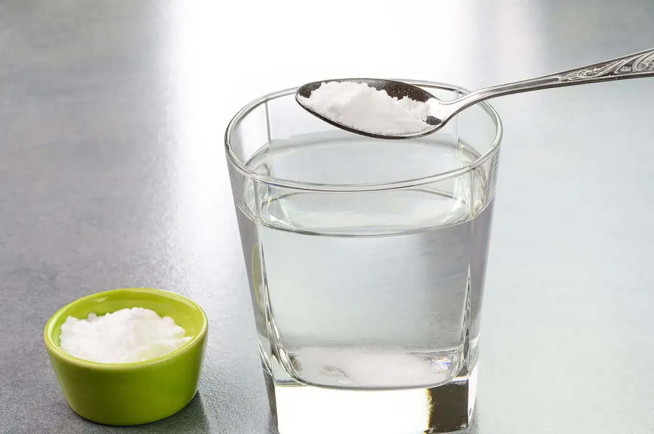 Une cuillère de bicarbonate de soude à boire est prête à être ajoutée à un verre d'eau. Une petite coupelle verte contient aussi du bicarbonate.