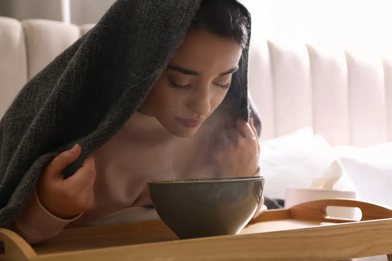 Jeune femme sous une couverture, faisant une inhalation pour son rhume. La vapeur s'&eacute;chappe d'un bol pos&eacute; sur un plateau.