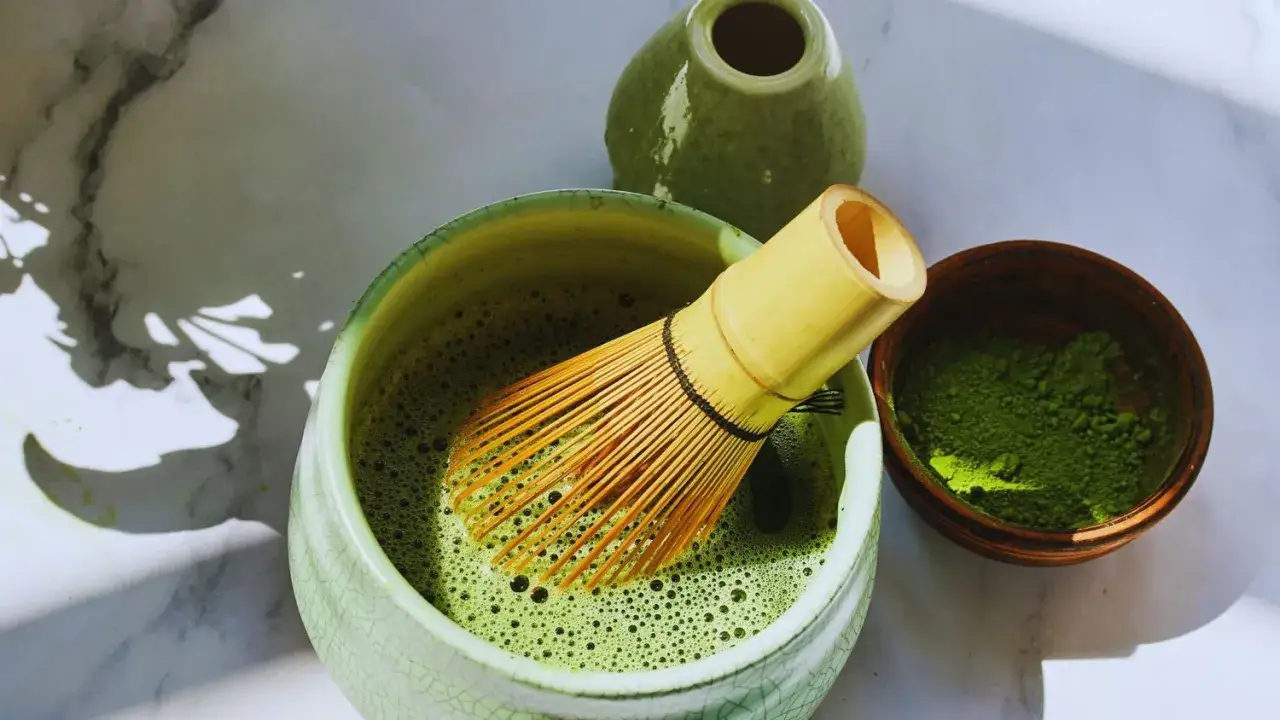 Préparation d'un thé matcha, révélant ses nombreux bienfaits. Un fouet en bambou dans un bol de matcha mousseux, à côté d'une petite coupelle de poudre verte.