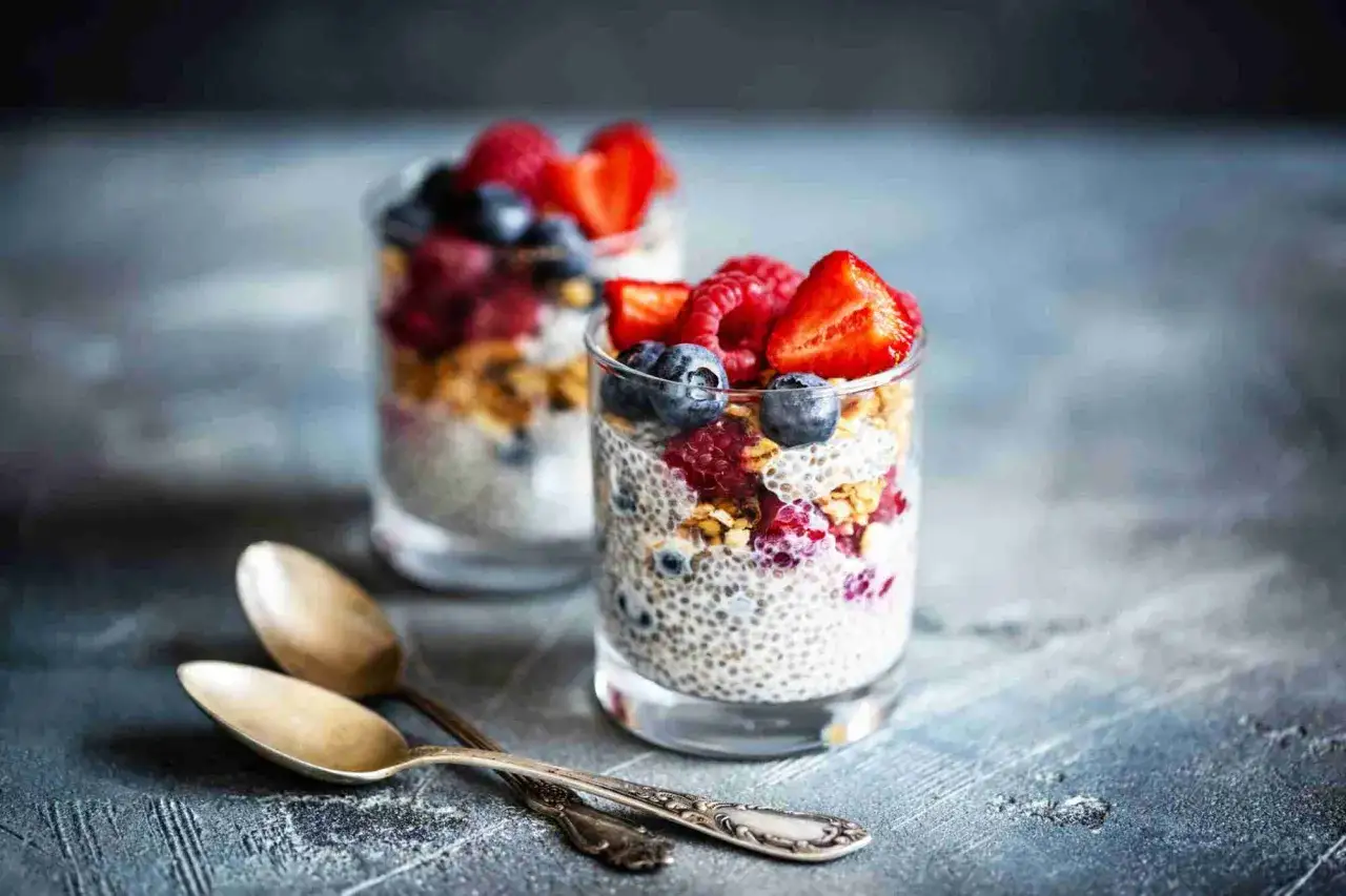Verres de pudding de graine de chia garnis de fruits rouges et de granola. Deux cuillères anciennes reposent sur la table.