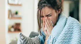 Femme enroulée dans une couverture bleue, se mouchant dans un mouchoir. Elle cherche un remède, peut-être un expectorant naturel.